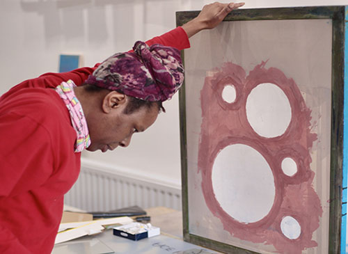 A woman is holding a piece of art work in progress. The work is made up of five circles and has a red outline. The lady has a purple headscarf on, and a red jumper.