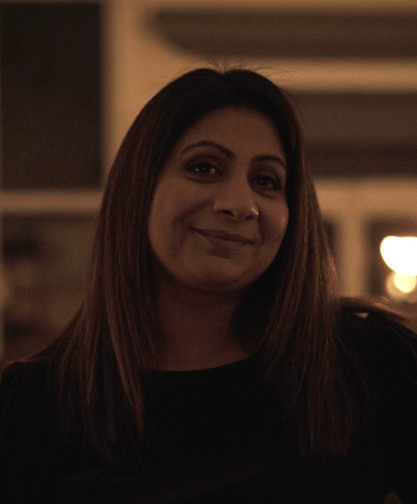 A woman smiles and looks directly at the camera. She has brown hair, brown eyes and olive skin. She wears a black top. Behind her the lights are blurred and a shelf is in view.