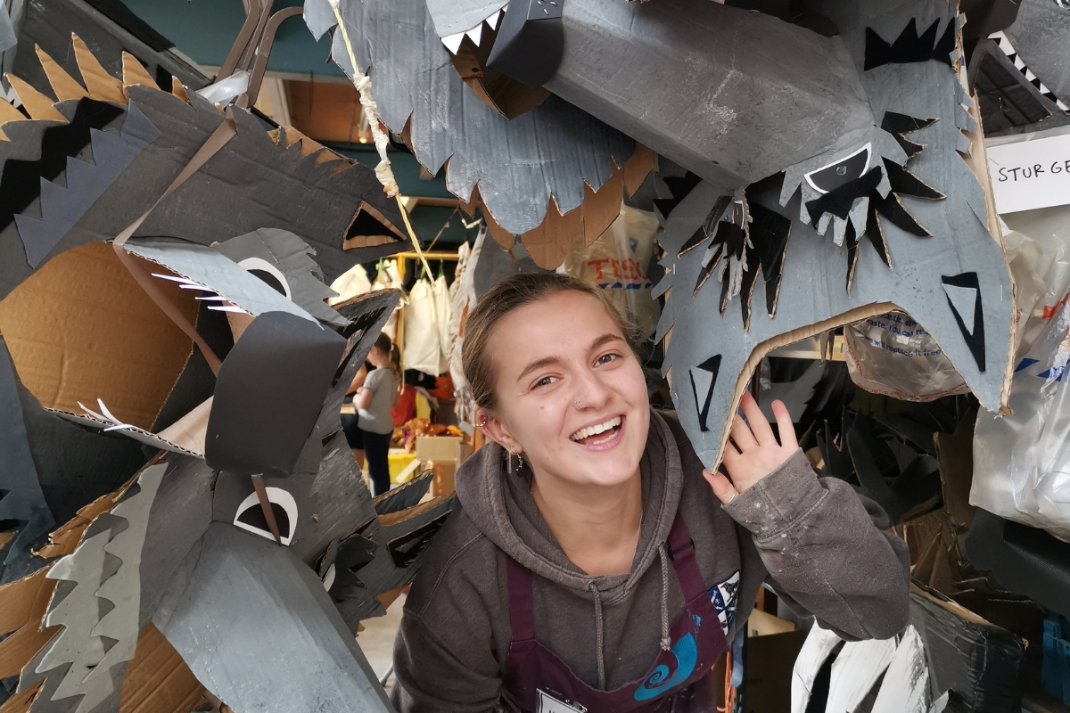 A white woman with blond hair is poking out through some grey cardboard statues, she is smiling.